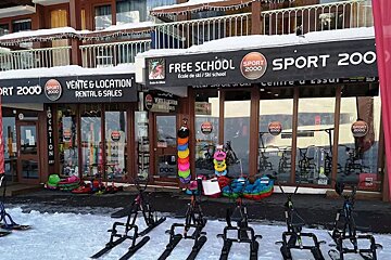 A snowy Sport 2000 ski rental and school with various winter sports equipment, including snow bikes, displayed outside in a mountain resort.
