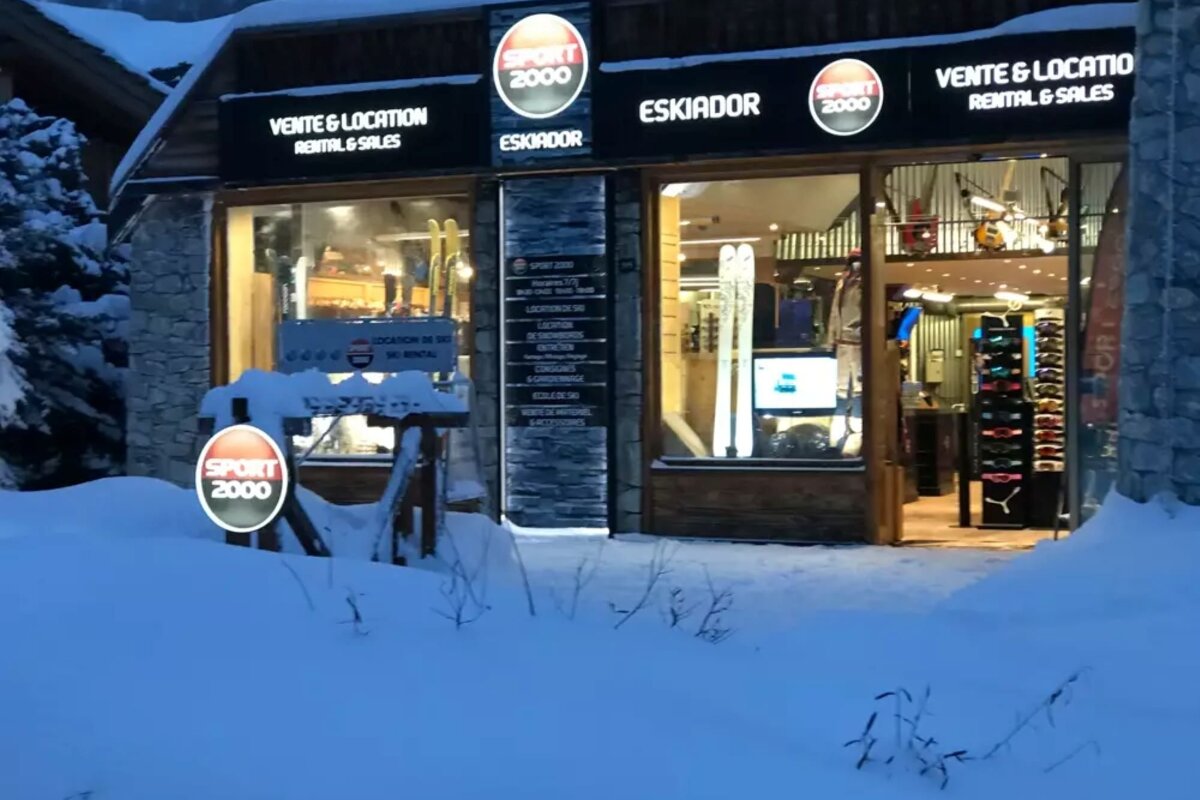 A snow-covered Sport 2000 ski rental shop, lit up at night, with skis and gear visible in the windows.