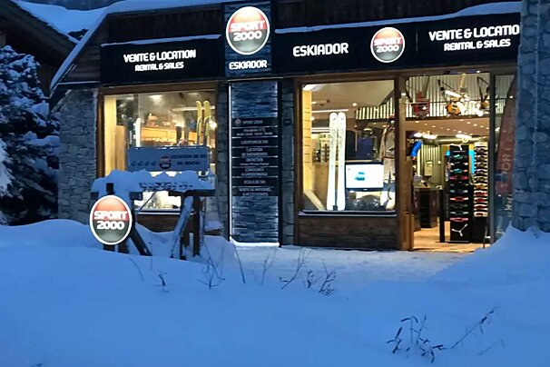 A snow-covered Sport 2000 ski rental shop, lit up at night, with skis and gear visible in the windows.