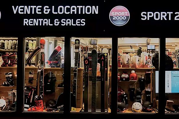 A brightly lit Sport 2000 shop window displays ski and winter sports equipment for rental and sales, including skis, boots, and helmets.