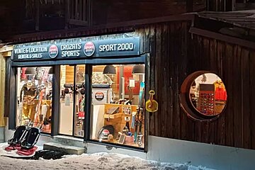 A brightly lit Sport 2000 ski/sports rental shop at night, with snow on the ground, showcasing winter gear in a wooden building.