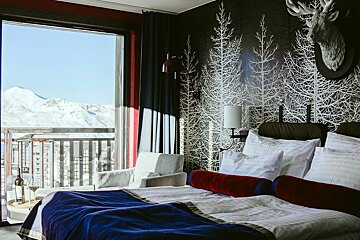 A stylish bedroom overlooking snowy mountains. Features a bed with blue/red accents, black tree wallpaper, a deer head, and wine on a balcony table.