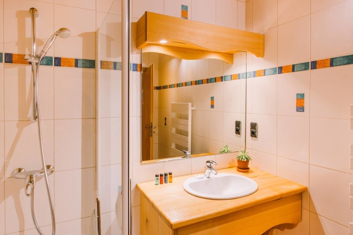A warm-toned bathroom with a shower, wooden vanity, sink, mirror, and white tiled walls accented with orange and blue decorative patterns.