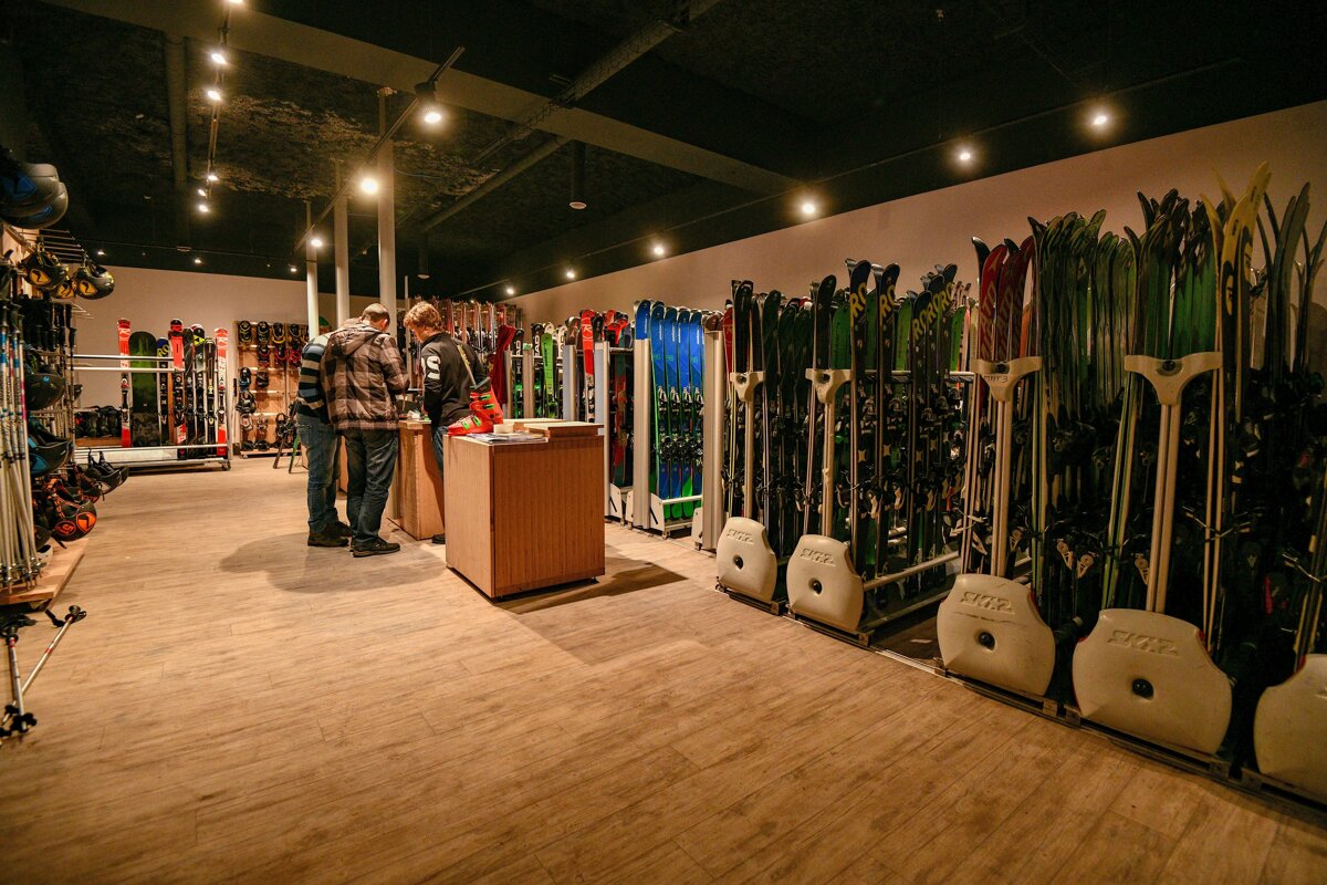 A ski and snowboard rental shop with rows of equipment. Three customers are at a counter, surrounded by gear under warm spotlights.