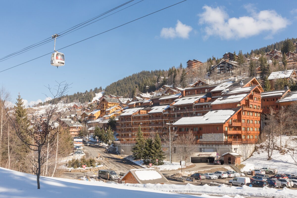 A cable car glides over a snowy alpine ski resort with wooden chalets under a bright blue sky.