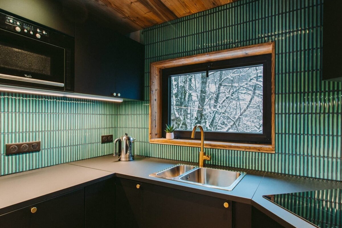 A modern kitchen featuring black cabinets, a green vertical tile backsplash, and a wooden ceiling. A window overlooks a snowy forest.