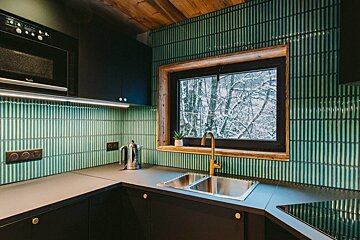 A modern kitchen featuring black cabinets, a green vertical tile backsplash, and a wooden ceiling. A window overlooks a snowy forest.