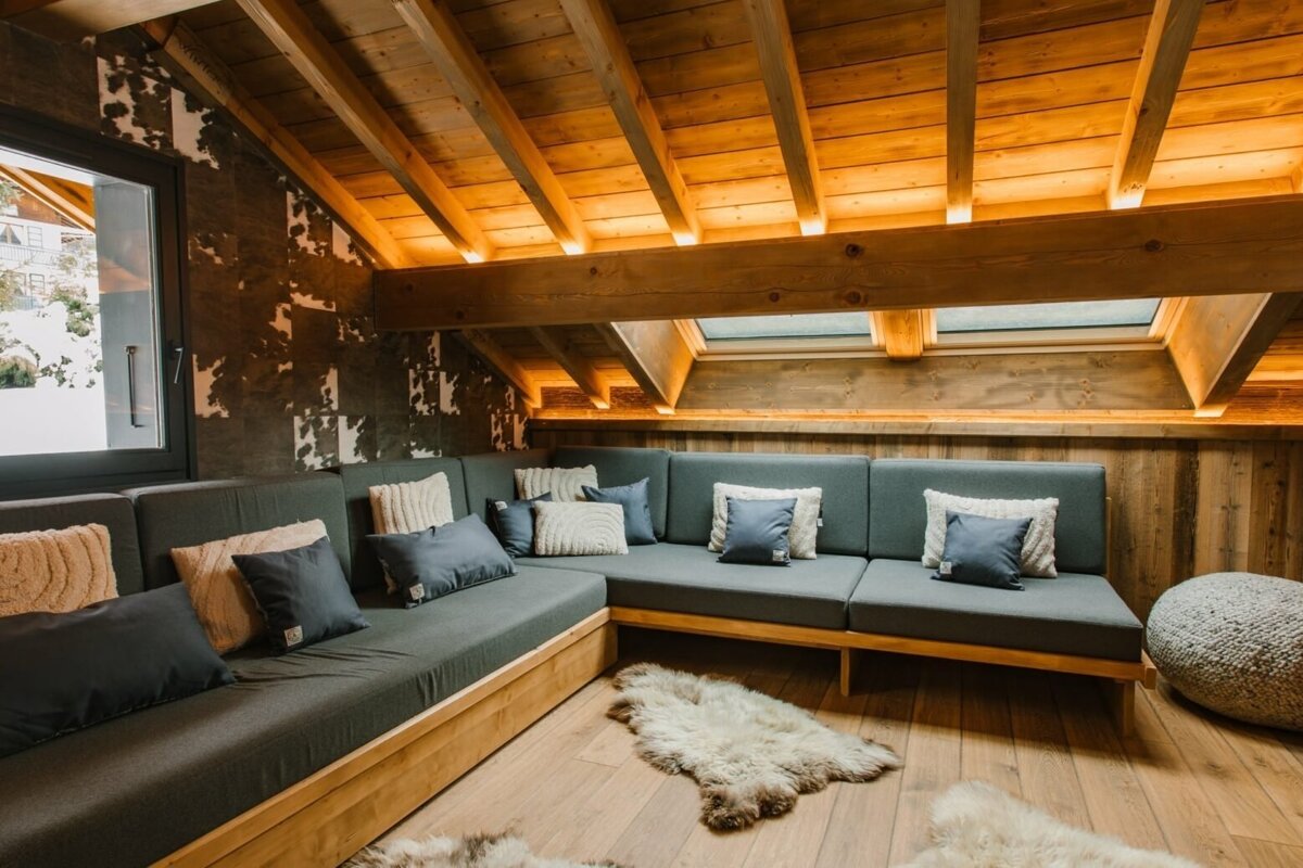 A warm, rustic attic lounge featuring wooden beams, ambient lighting, an L-shaped gray sofa with pillows, and soft fur rugs on a light wood floor.