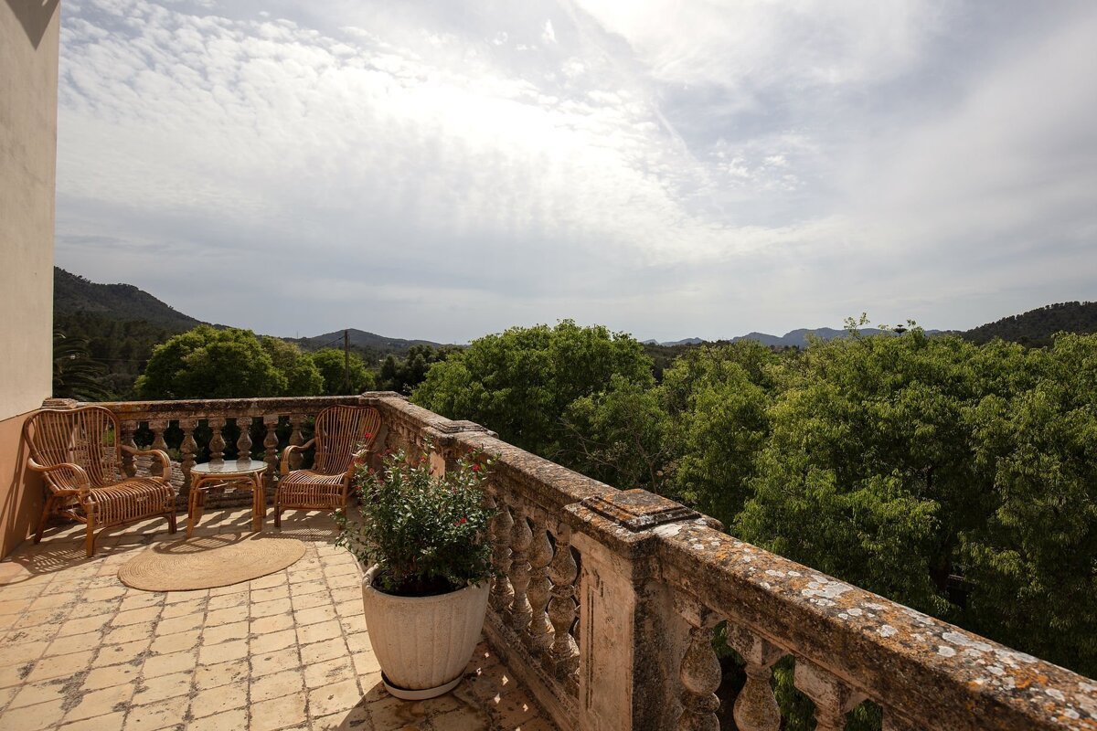 An outdoor balcony with wicker chairs and a table, featuring a stone railing and a stunning vista of a lush green forest, distant mountains, and a cloudy sky.