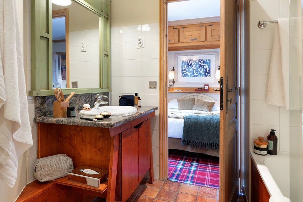 Rustic bathroom with a wooden vanity and green-framed mirror, looking into a cozy bedroom with a plaid rug.