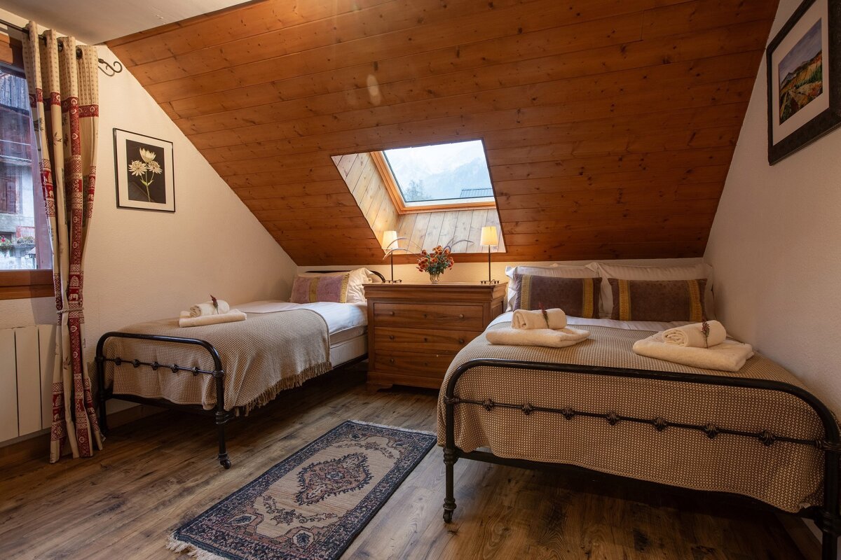 A rustic attic bedroom with a sloped wooden ceiling, two single beds, and a skylight. A dresser sits between the beds, illuminated by lamps, on a wooden floor with a rug.