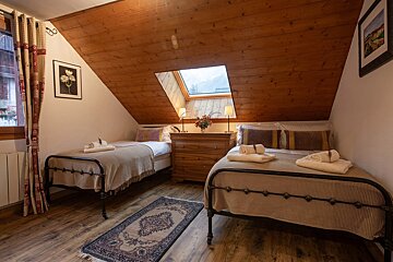 A rustic attic bedroom with a sloped wooden ceiling, two single beds, and a skylight. A dresser sits between the beds, illuminated by lamps, on a wooden floor with a rug.