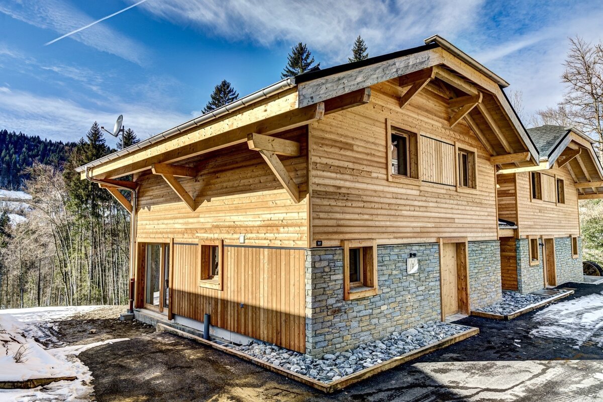 A modern wood and stone chalet-style house with exposed beams, nestled in a snowy, forested landscape under a bright blue sky.