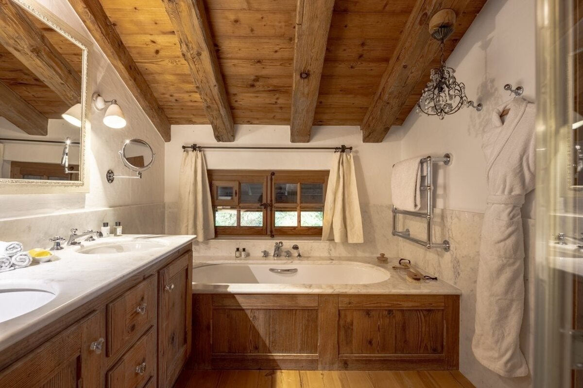 Rustic bathroom with exposed wooden beams, a built-in bathtub, double marble vanity, and a window. A cozy, warm-toned space.