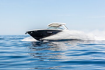 A sleek motorboat speeds across calm blue water, kicking up a large white spray under a clear sky.