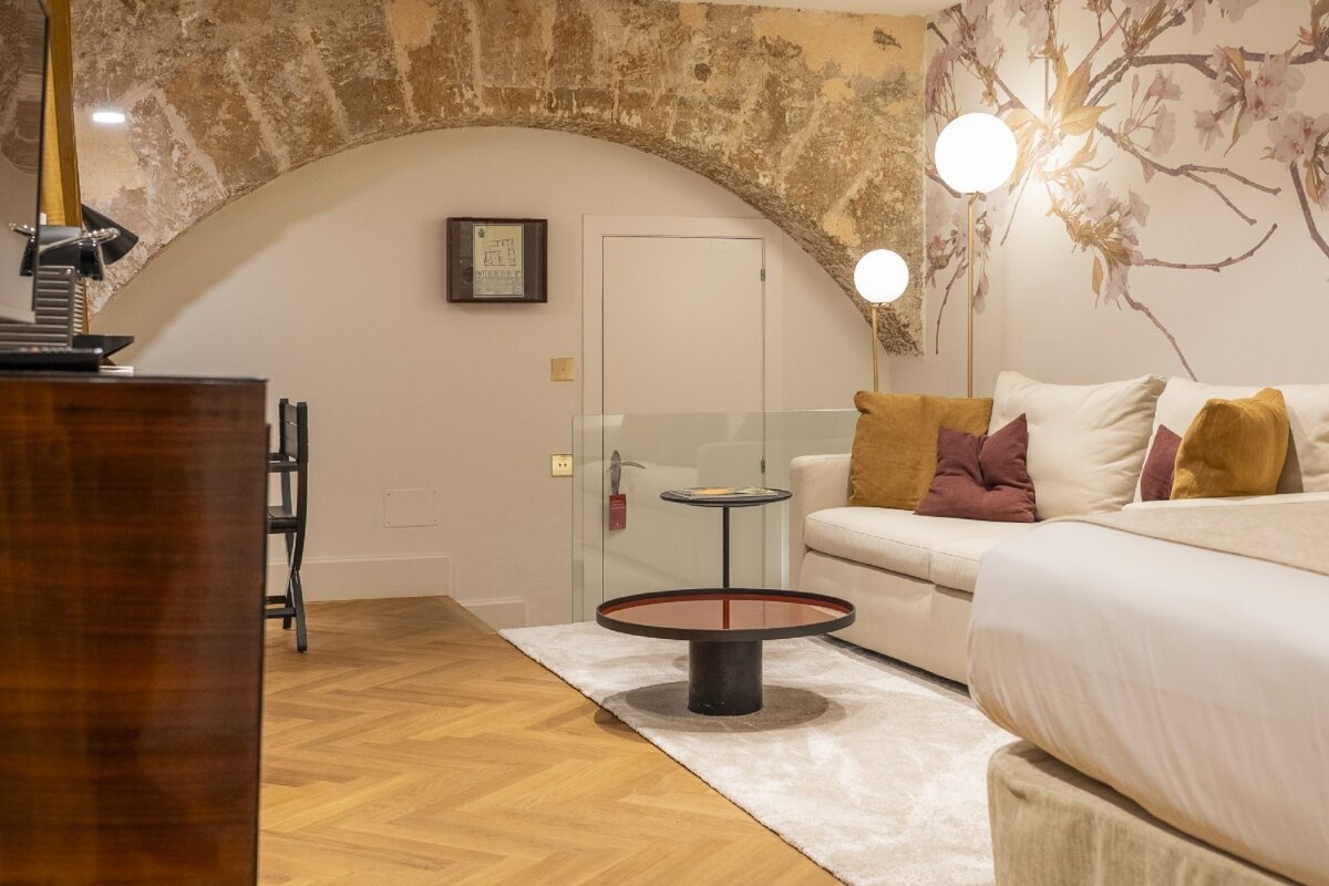 A chic room blends ancient stone arches with modern design. It features a sofa, bed, floral wallpaper, herringbone floor, and stylish lighting.