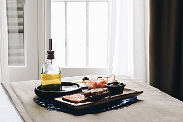 An appetizer tray with olive oil, bread, prosciutto, olives, and cheese sits by a bright window with white curtains.