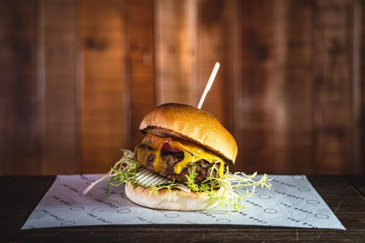 A juicy burger with melted cheese, lettuce, and a skewer, served on branded paper against a rustic wooden background.