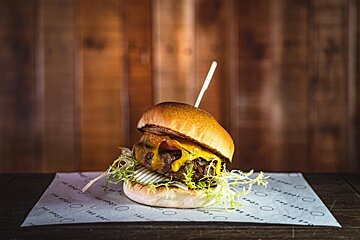 A juicy burger with melted cheese, lettuce, and a skewer, served on branded paper against a rustic wooden background.