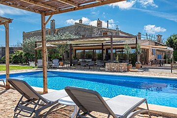 A serene blue swimming pool with loungers in the foreground, set against a historic stone building and a shaded outdoor dining area on a sunny day.