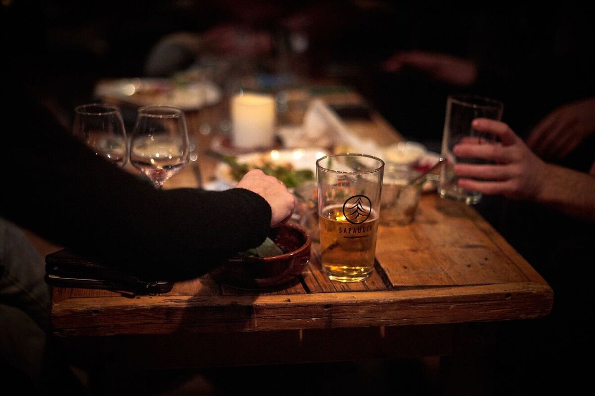 A dimly lit wooden table with a beer, wine glasses, and hands reaching for food, suggesting a cozy evening gathering.