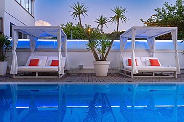 A serene hotel pool area at dusk with two white cabanas, orange pillows, a large plant, and blue ambient lighting reflecting in the water.