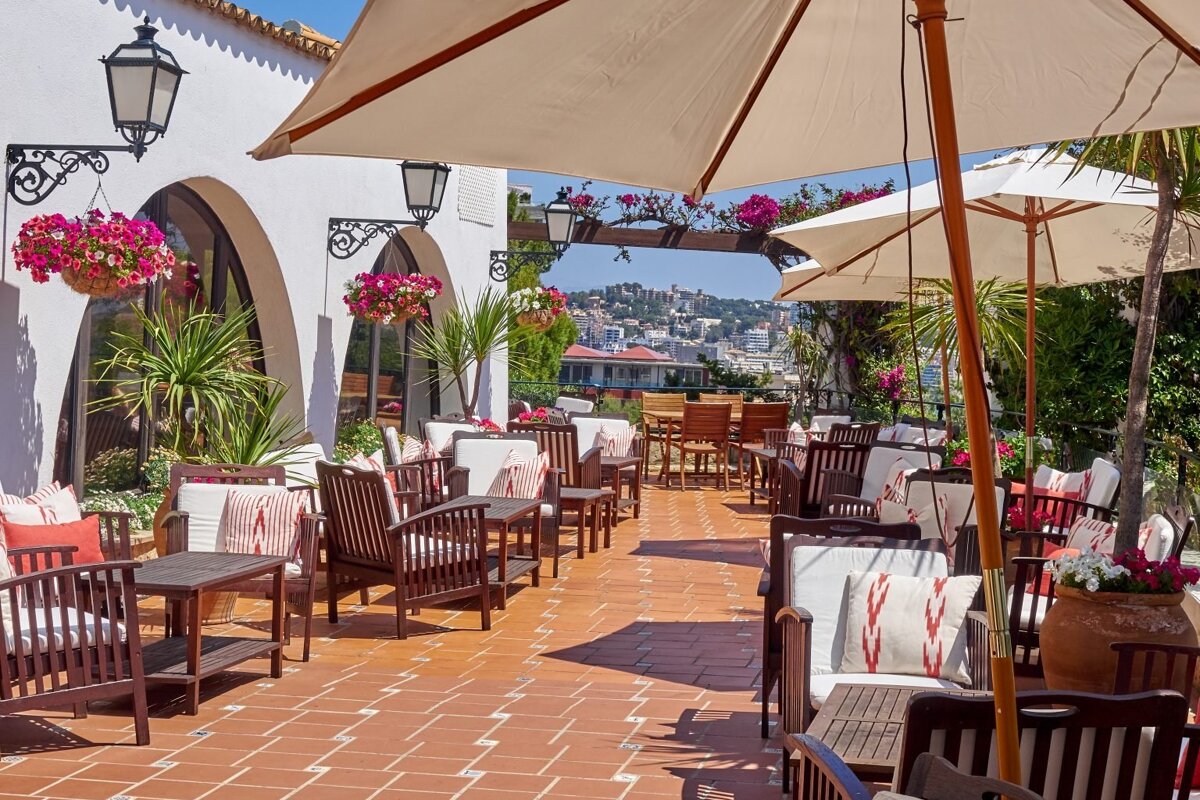 A beautiful Mediterranean-style outdoor terrace with wooden seating, white umbrellas, and vibrant flowers, offering a scenic city view.