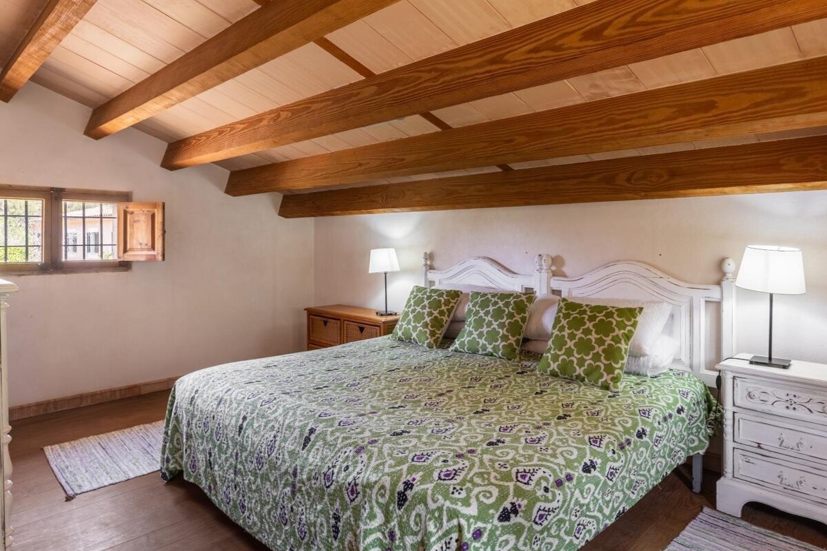 Cozy bedroom with white walls, wooden beamed ceiling, a bed with green patterned bedding, white headboard, and a window with wooden shutters.