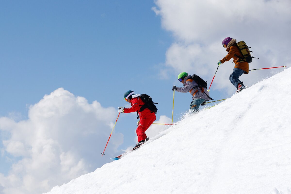 Three skiers in colorful gear carve down a snowy mountain slope under a bright blue sky with fluffy clouds.