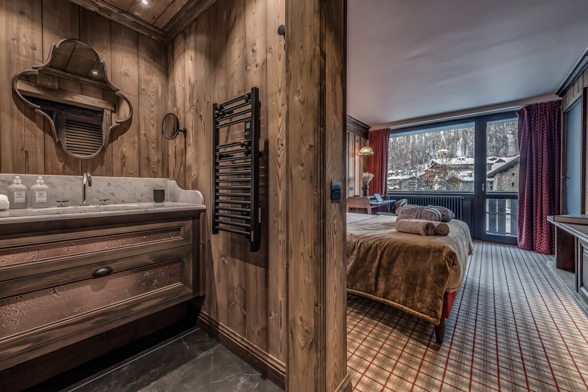 A cozy, wood-paneled chalet room featuring a rustic bathroom vanity, heated towel rack, and a bedroom with a large window overlooking a snowy village.
