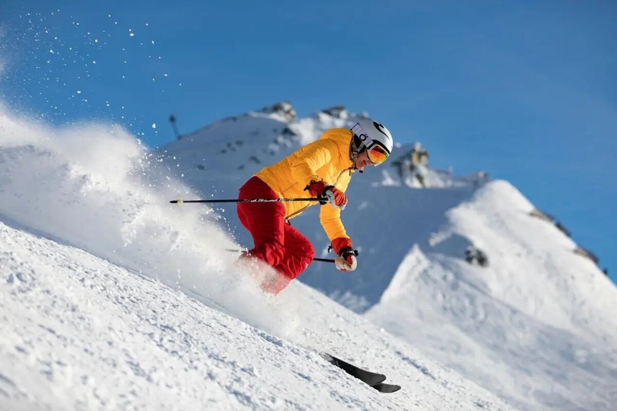 A skier in a yellow jacket and red pants carves down a snowy mountain slope, kicking up powder under a bright blue sky with distant peaks.