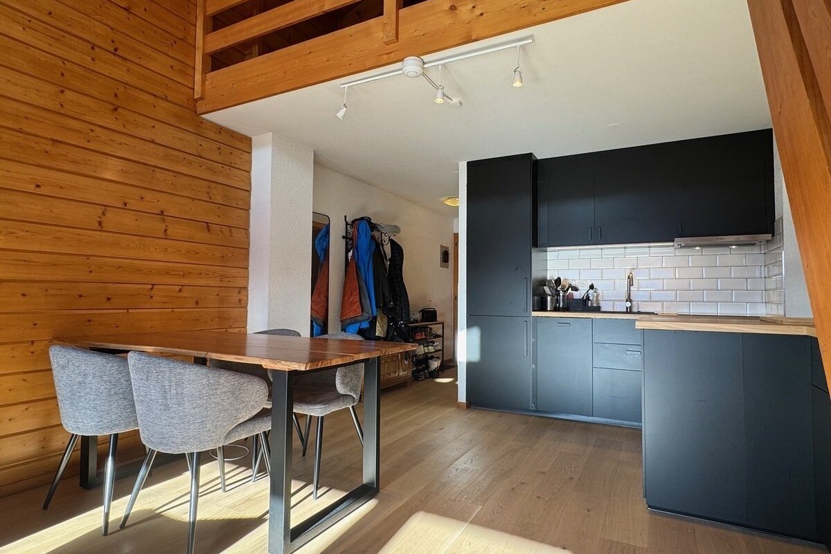Open-plan room with wood-paneled walls, a dining table with chairs, a modern black kitchen, and a wooden floor, with a loft visible above.