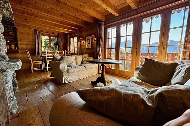Warm, rustic wooden chalet interior with cozy sofas, dining space, and large windows looking out to stunning mountain views.