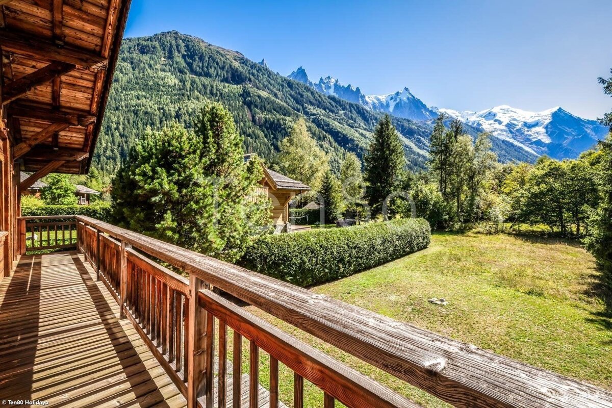 A wooden balcony overlooks a green field and a hedge, with lush forested mountains and majestic snow-capped peaks under a bright blue sky.