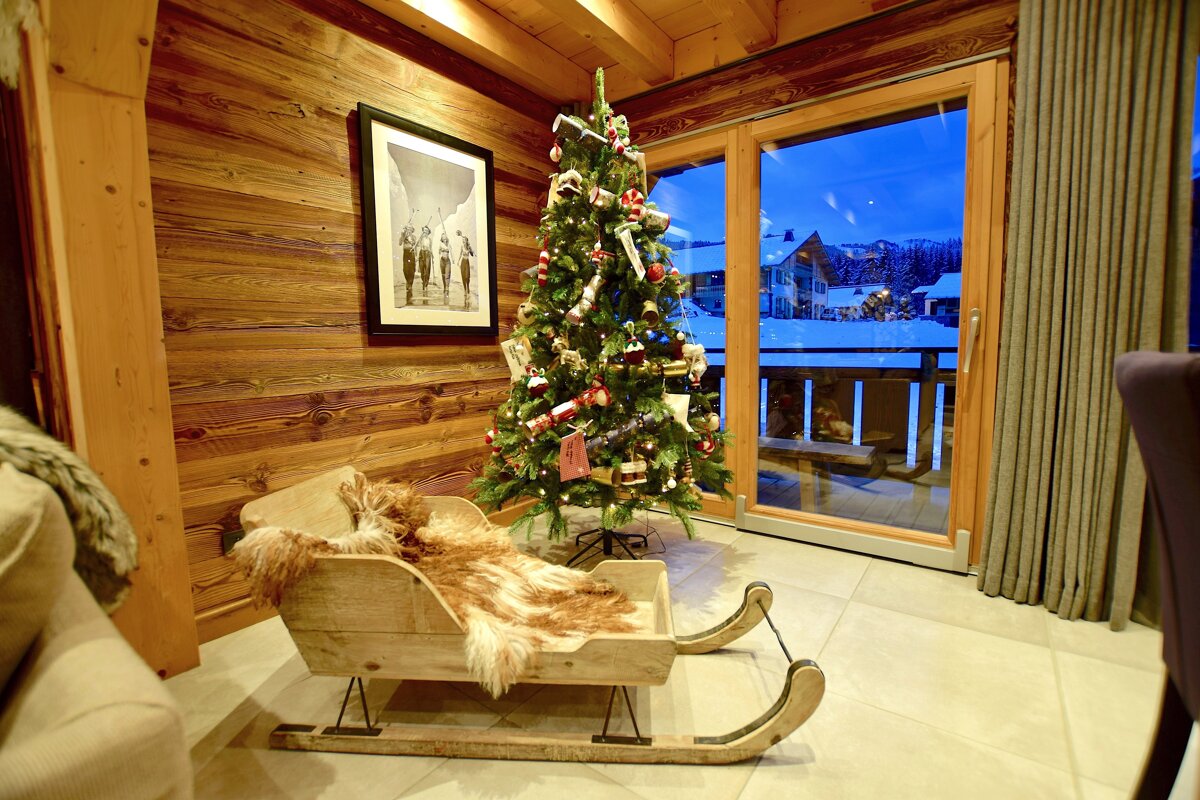 A cozy wooden chalet with a festive Christmas tree, fur-lined sleigh, and a window revealing a snowy mountain village at blue hour.
