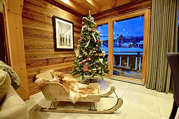 A cozy wooden chalet with a festive Christmas tree, fur-lined sleigh, and a window revealing a snowy mountain village at blue hour.