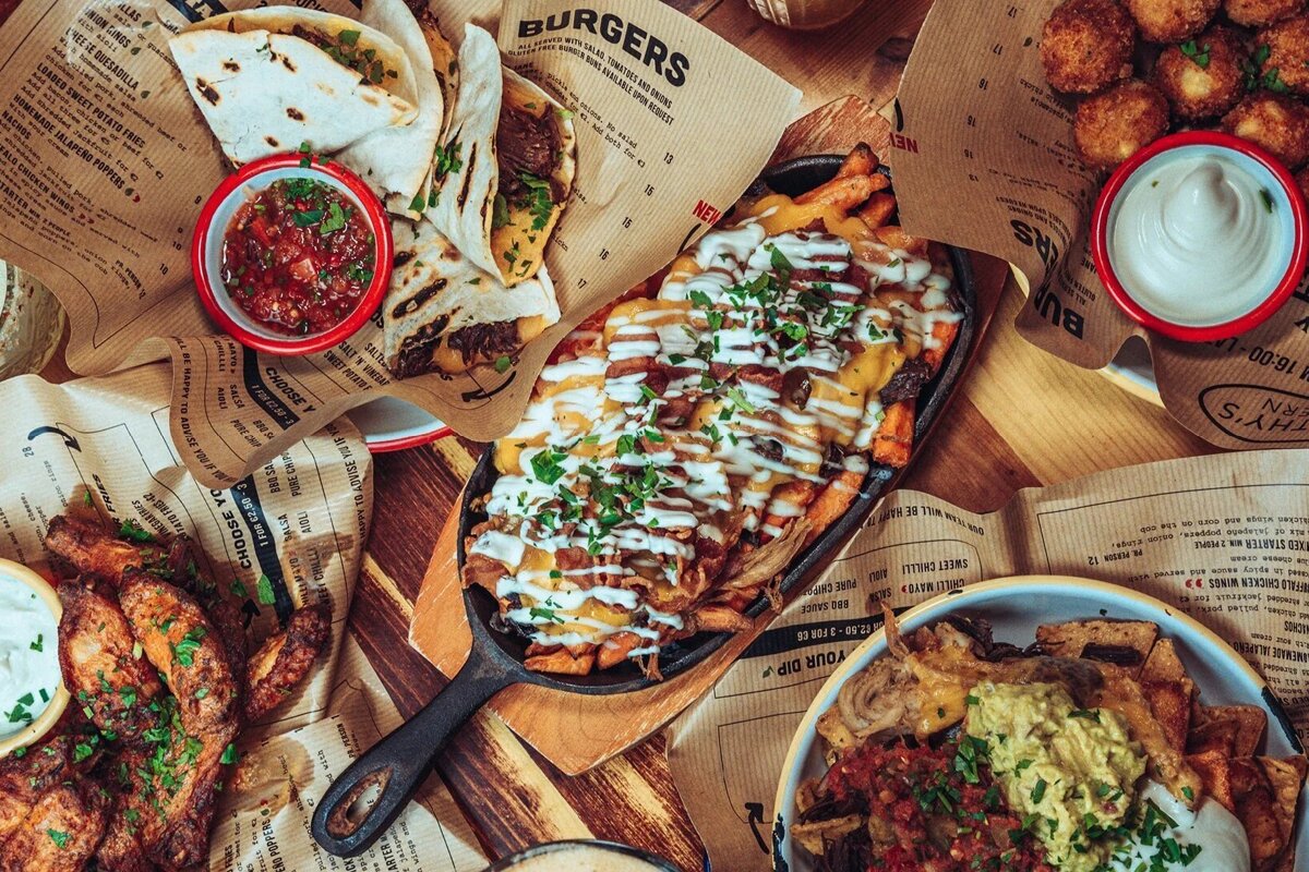 A vibrant overhead view of a table laden with comfort foods like loaded fries, tacos, nachos, wings, and dips, served on menu paper.