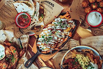 A vibrant overhead view of a table laden with comfort foods like loaded fries, tacos, nachos, wings, and dips, served on menu paper.