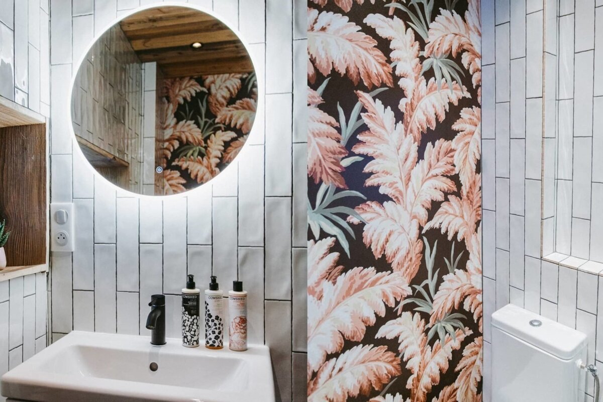Modern bathroom: white tiles, backlit round mirror, sink, and bold leafy floral wallpaper.