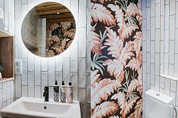 Modern bathroom: white tiles, backlit round mirror, sink, and bold leafy floral wallpaper.