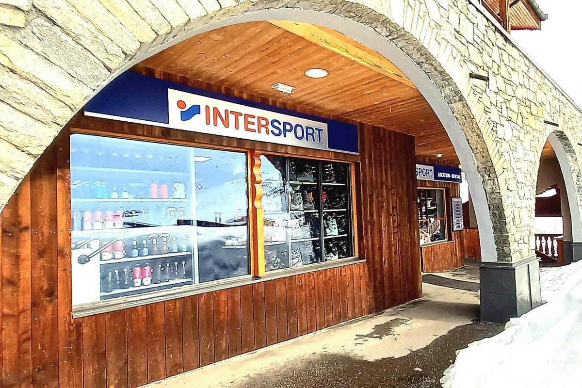 An Intersport shop with wooden walls under a stone archway, displaying items through large windows. Snow covers the ground outside.