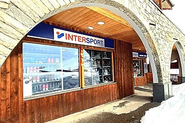 An Intersport shop with wooden walls under a stone archway, displaying items through large windows. Snow covers the ground outside.