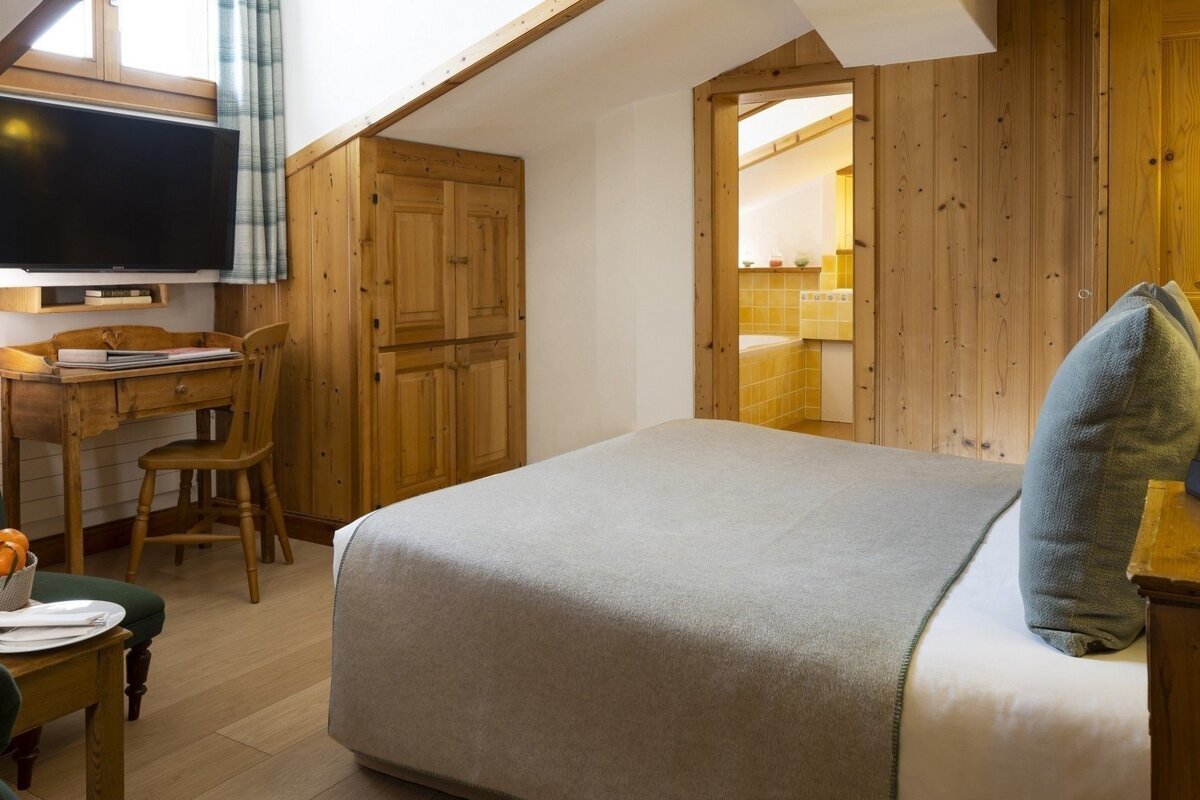A cozy, wood-paneled bedroom with a light grey bed, desk, TV, and a glimpse of a yellow-tiled bathroom through an open doorway.