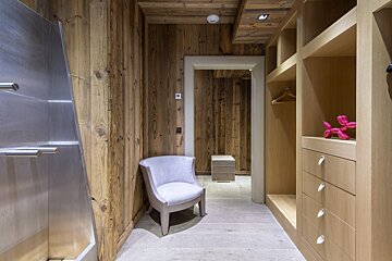 A rustic modern walk-in closet featuring warm wood paneling, a sleek stainless steel door, a grey armchair, and light wood shelves with a pink balloon dog.