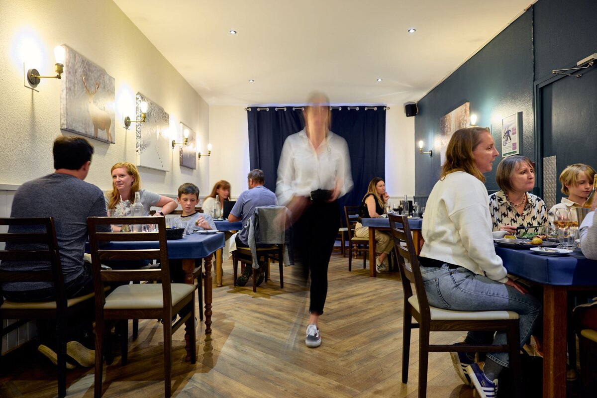 A restaurant interior with people dining at tables, a blurred staff member walks through the middle. Blue tablecloths & light walls.