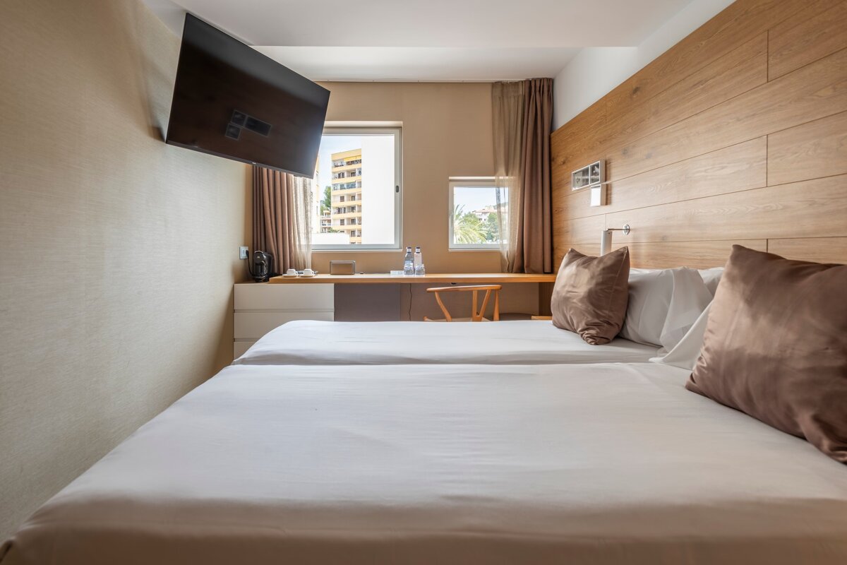 Modern hotel room with two single beds, a wooden headboard, wall-mounted TV, and a desk under windows offering an outside view.