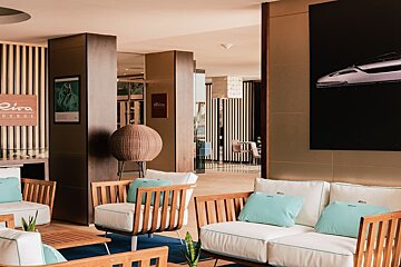 Stylish Riva Lounge featuring modern wooden seating with white and blue cushions, a large yacht artwork, and contemporary decor.