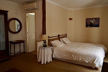A cream-colored bedroom with a bed, white linens, a wooden mirror, small tables, a lamp, an AC, and a painting, creating a rustic yet cozy feel.