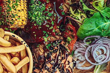 A hearty BBQ meal with saucy ribs, grilled corn on the cob, fries, fresh mixed greens, sliced tomatoes, red onion, and coleslaw.