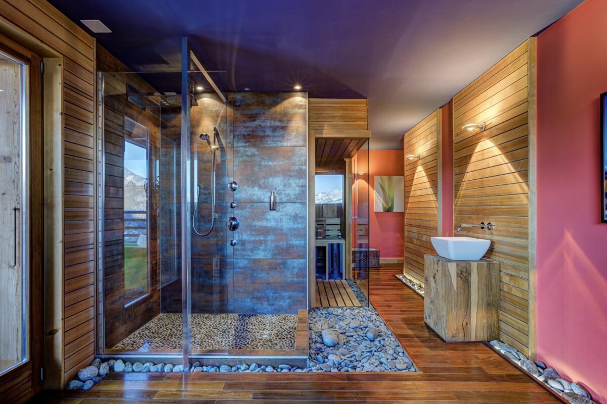 A stylish, rustic-modern bathroom featuring a glass shower with pebble floor, wooden walls/accents, and a sink on a wood block. Bold coral and purple colors.